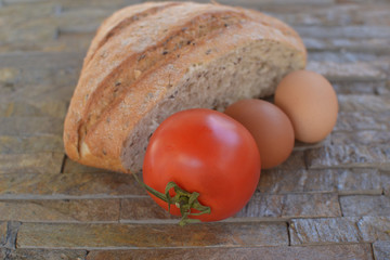 half bread of black flour with seeds red tomatoe with dried green stem and two eggs on a plate of...