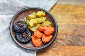 clay dish with delicious baked onions, carrots and potatoes stands on a wooden stand and a linen napkin