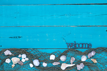 Welcome sign hanging on antique rustic teal blue wood sign with seashells and fish netting border; wooden background with painted copy space