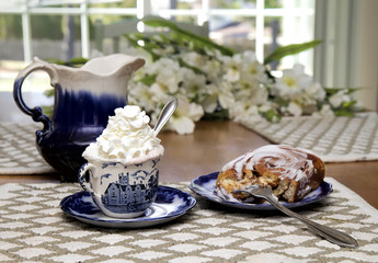 Morning breakfast snack with hot chocolate loaded with whipped cream and large cinnamon danish.  Served on antique flow blue dishes at kitchen table.  Flowers in backround.