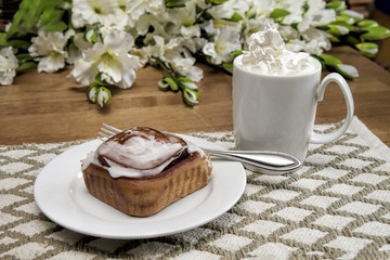 Morning breakfast snack with hot chocolate in large white mug loaded with whipped cream and large cinnamon danish.  Flowers on table.