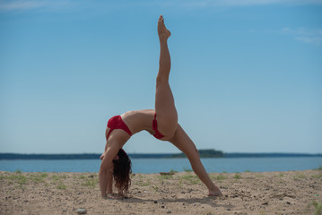 Fototapeta na wymiar Young Woman Relaxing and Sunbathing At Beach. Beautiful woman in red bikini doing yoga while standing on bridge pose with sea view on background