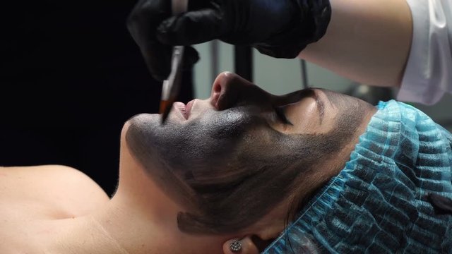 Preparation of face for cosmetic procedure. Cosmetologist applies carbon compound on skin with brush. Carbon peeling. Shooting close-ups