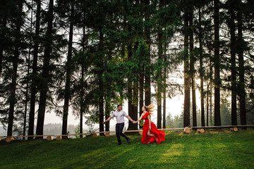 Beautiful happy couple in love is running by the hand along the green summer forest during the sunset.