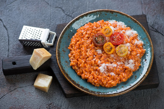 Plate of risotto with tomatoes and parmesan on a black wooden serving board, grey cracked asphalt background, studio shot
