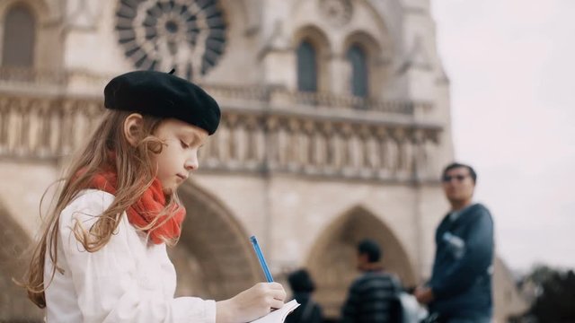 Little cute girl in beret and red scarf sitting near the Notre Dame and drawing the sketch in Paris, France.
