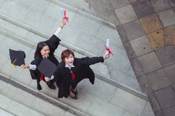 Portrait of Two happy graduated young girls in graduation gowns holding diplomas and smile together in city form top view.