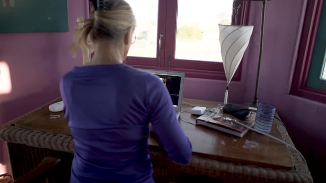Slow steadicam orbit to the left of a woman on a laptop computer in a bedroom at a desk.