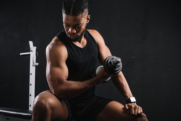 Fototapeta na wymiar young african american sportsman exercising with dumbbell and looking at biceps