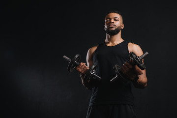 Fototapeta premium low angle view of young african american sportsman holding dumbbells and looking at camera isolated on black