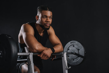 Fototapeta na wymiar muscular young african american sportsman leaning at barbell and looking away on black