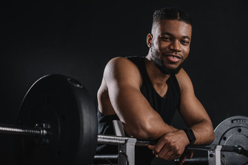 muscular young african american sportsman leaning at barbell and smiling at camera on black