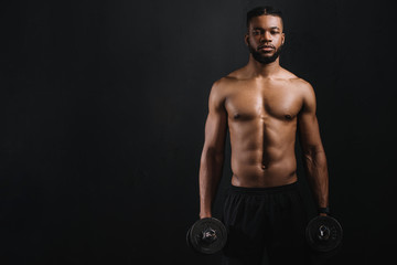 Fototapeta na wymiar muscular shirtless african american man holding dumbbells and looking at camera isolated on black