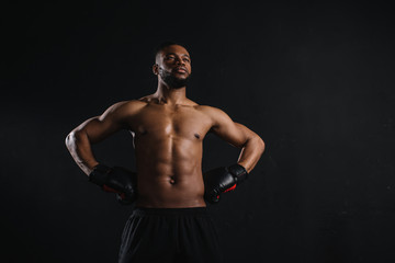 Fototapeta na wymiar muscular shirtless african american man in boxing gloves standing with hands on waist and looking away isolated on black