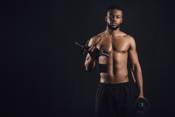 Fototapeta na wymiar muscular shirtless african american sportsman exercising with dumbbells and looking at camera isolated on black