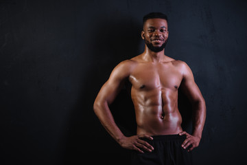 Fototapeta na wymiar shirtless muscular young african american man standing with hands on waist and smiling at camera on black