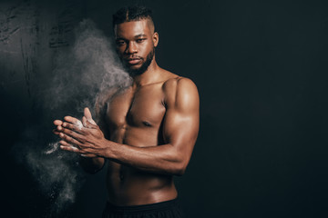 Fototapeta na wymiar young bare-chested african american sportsman applying talcum powder and looking at camera on black