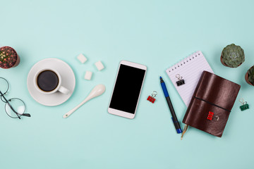 Business flatlay with smartphone with black copyspace screen, cup of coffee, succulents and cactuses and other business accessories. Mint color background. Top view
