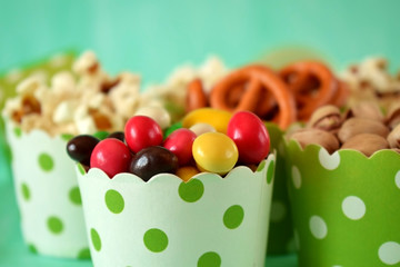 Popcorn, multicoloured drops, pretzels with salt and pistachio nuts in paper cups on green background. Snacks assortment