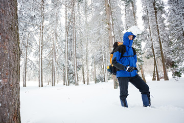 Fototapeta na wymiar Young male mountain climber in blue and black clothing with hood hiking toward camera through deep powder snow in pine forest. Wide angle view.