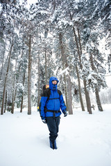Fototapeta na wymiar Young male mountain climber in blue and black clothing with hood hiking toward camera through deep powder snow in pine forest. Wide angle view.