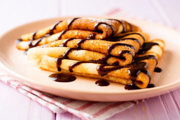 Crepes with chocolate syrup on purple wooden background.