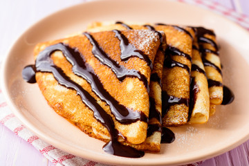 Crepes with chocolate syrup on purple wooden background.