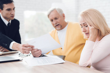 A couple of old people came to see a realtor. An old man in yellow cardigan and woman in a sweater are sitting at table.