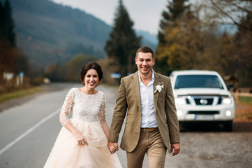 Newlyweds are walking, the green hill with pines and big white car on background