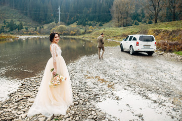 The newlywed are walking on the rocky river coast, white car and mountains on background