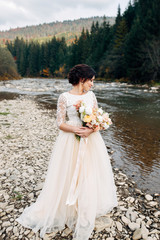 Beautiful dark-haired bride in beige dress with wedding bouquet on the riverside, mountains and pines on background