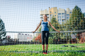 Beautiful stylish woman in a blue shirt and leggings stands near a football goal at the stadium at sunset. Beautiful sunlight. A girl with a perfect figure and in great shape.