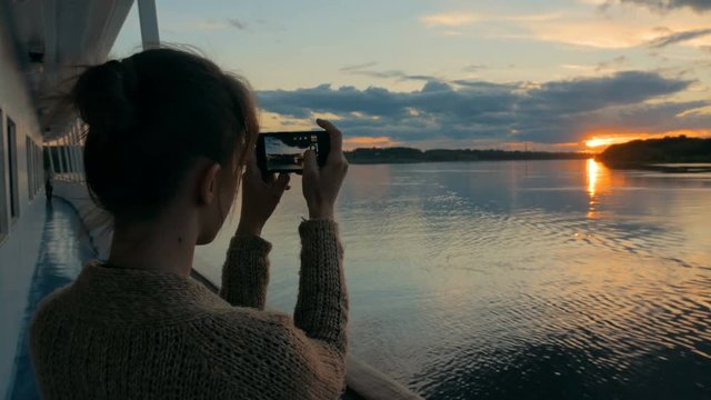 Woman silhouette taking photo of beautiful sunset with smartphone on deck of cruise ship. Sunset light, golden hour. Photography, nature and journey concept