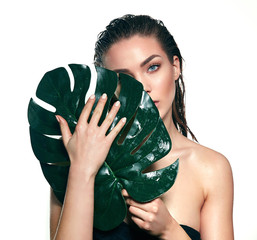 Obraz premium A beautiful young girl in the studio on a white background with wet skin and wet hair holds a large green tropical leaf in hands and covers a part of her face.