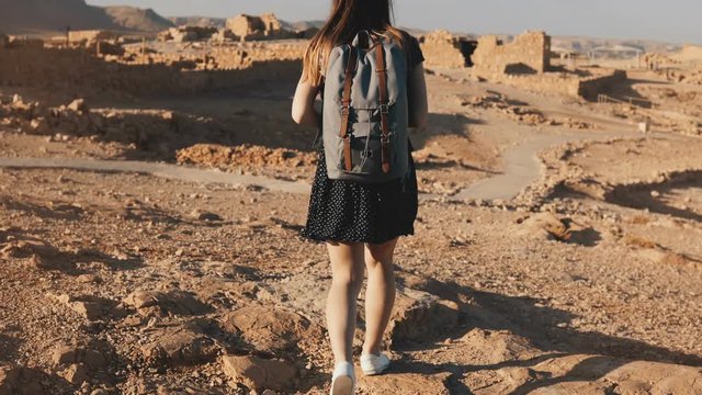 Woman with backpack explores ancient desert ruins. Beautiful European tourist walks on rocks and sand. Masada Israel 4K.