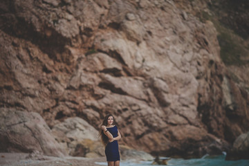 A young beautiful girl in a tight dark blue dress dreams on the beach. Blurred rocks and cliffs on background. Horizontal orientation. Copy space