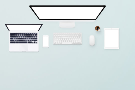 Top view of a light blue desk with a laptop, desktop computer, smartphone, tablet, keyboard, mouse and coffee. Devices with isolated screen on blue table background