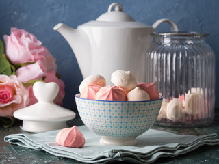 Pink and white meringues in ceramic china bowl on blue background with roses and teapot