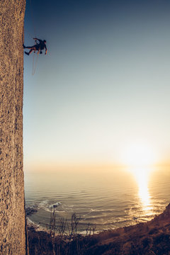 Man abseiling down mountain cliff overlooking sea at sunset
