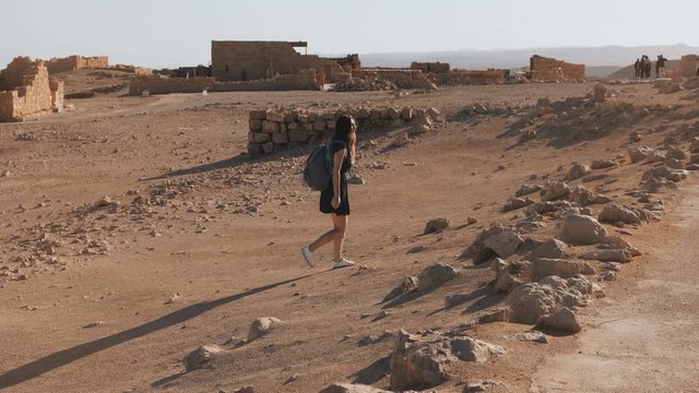 Girl with backpack explores desert ruins. Pretty European female traveler walks among ancient scenery. Masada Israel 4K.
