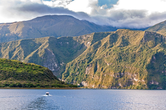 View of the Cuicocha lake and crater, with a small touristic boat in the water, on a sunny and cloudy afternoon.