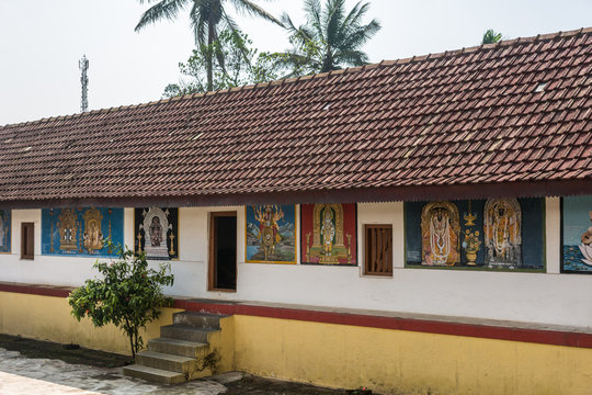 Madikeri, India - October 31, 2013: Shree Omkareshwara Temple inside courtyard. Colroful Hindu Religious wall paintings on perimeter building with large red roof. Silver sky and some green trees.