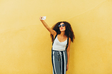 Portrait of a chic tourist taking a selfie in front of a yellow wall.