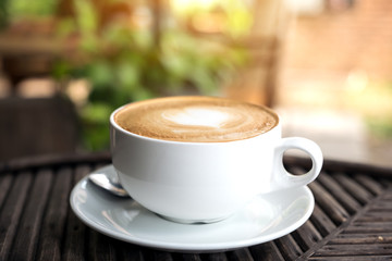 A white cup of hot latte coffee with a heart latte art on vintage wooden table with blur green nature background