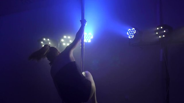 A flexible sexy girl in striped stripes is dancing a sensual dance on a pole in the dark under the lights of multi-colored searchlights. Pole dance. Slow motion.
