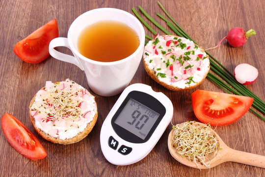 Glucose meter with result of sugar level, freshly sandwich with vegetables and hot tea, healthy nutrition
