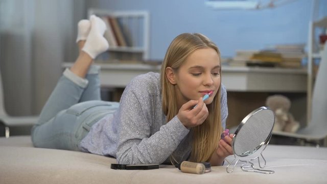 High-school girl applying her first pink lip gloss admiring herself, adolescence