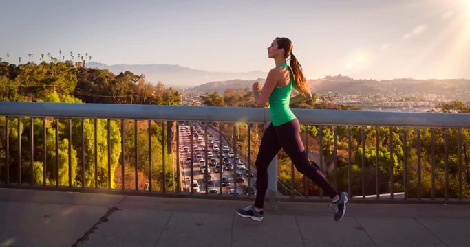 Athletic Woman Working Out. Jogging across a bridge. Trees, Sun and city can be seen in the background. Slow Motion.