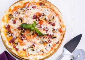 Hot homemade pizza with sausage, mushrooms, mozzarella cheese and basil on wooden board, copy space. Pizza cutter and spatula server. Italian traditional cuisine. Italian food. Top view, flat lay