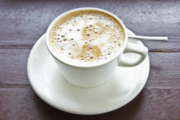 Coffee latte with milky foam and bubbles in a white porcelain cup on a saucer on a wooden brown table.